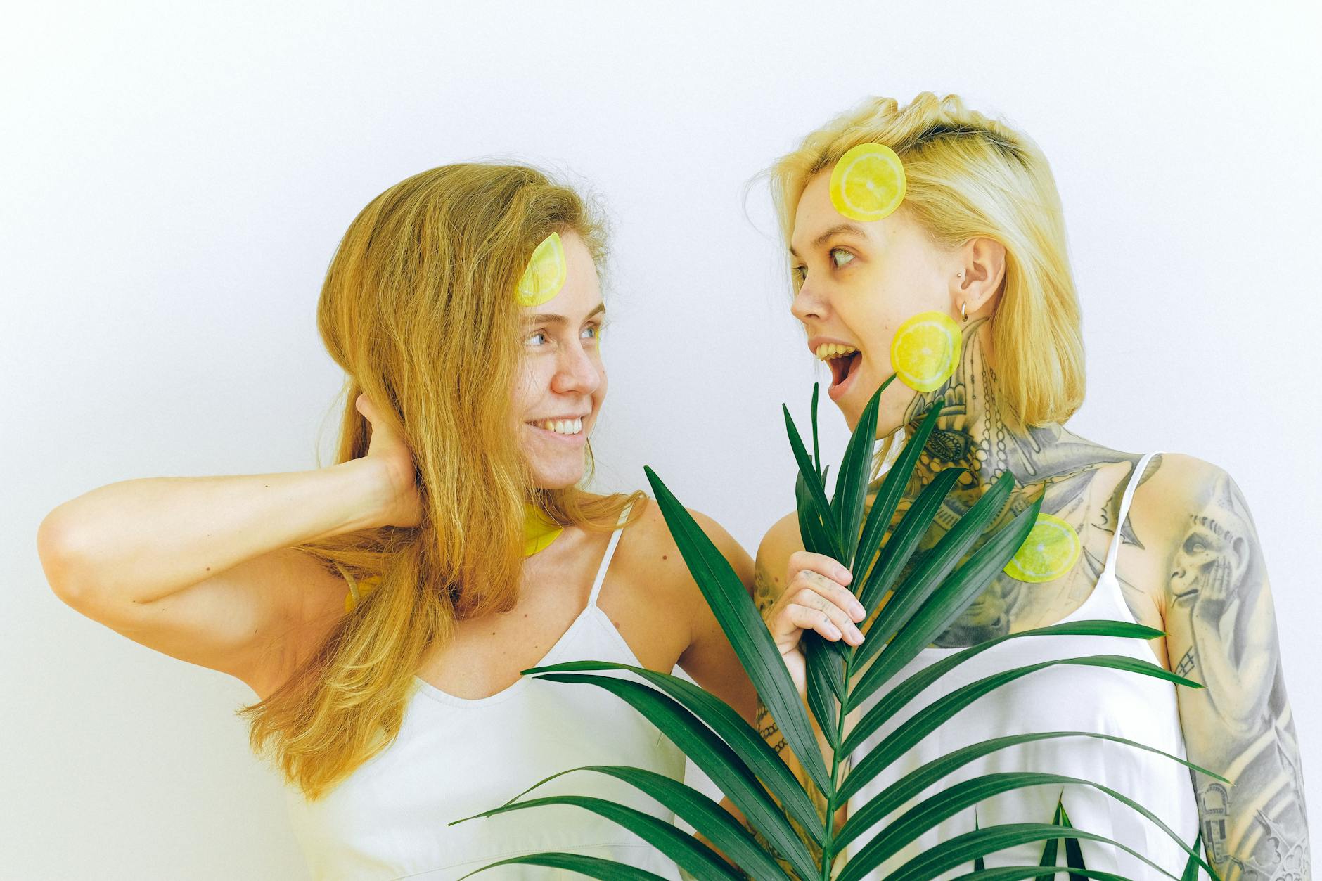 Two women smiling with lemons and tropical plants, expressing genuine joy and comfort together indoors
